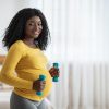 Happy pregnant black woman lifting dumbbells, exercising at home