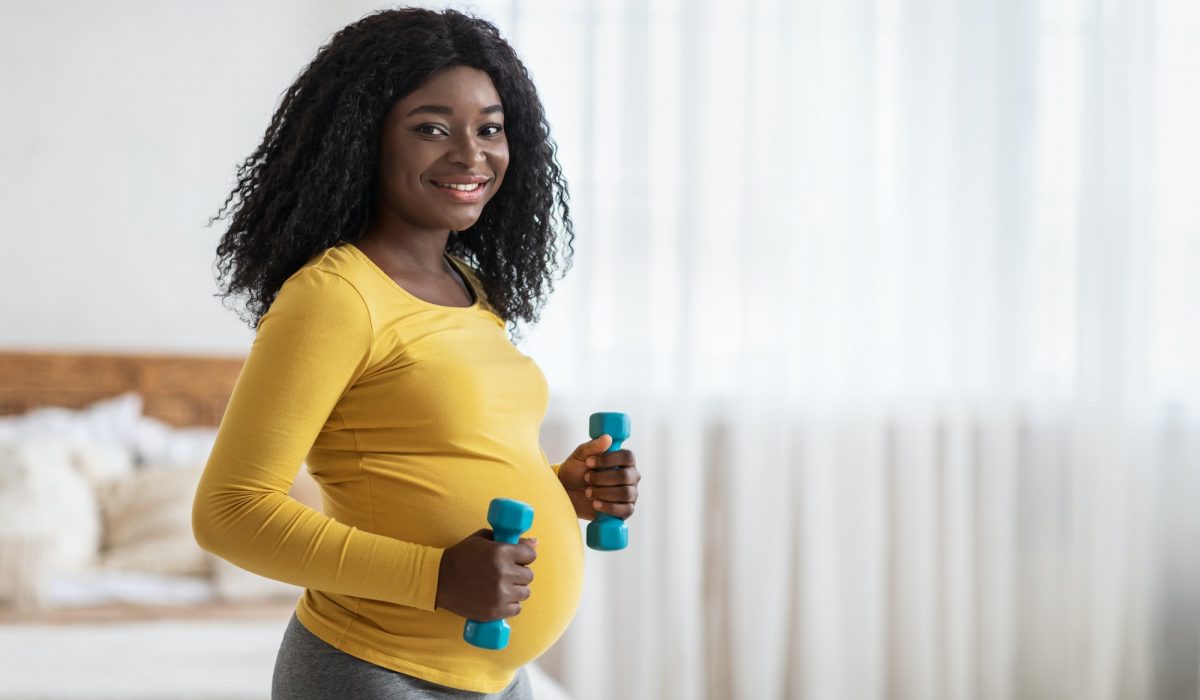 Happy pregnant black woman lifting dumbbells, exercising at home